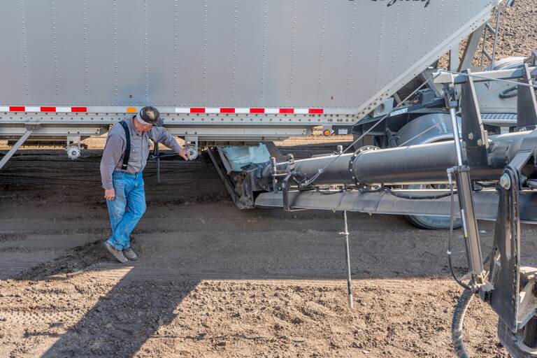 A farmer works the crank beneath a fertilizer hopper trailer as it loads into an auger on a tilled prairie field